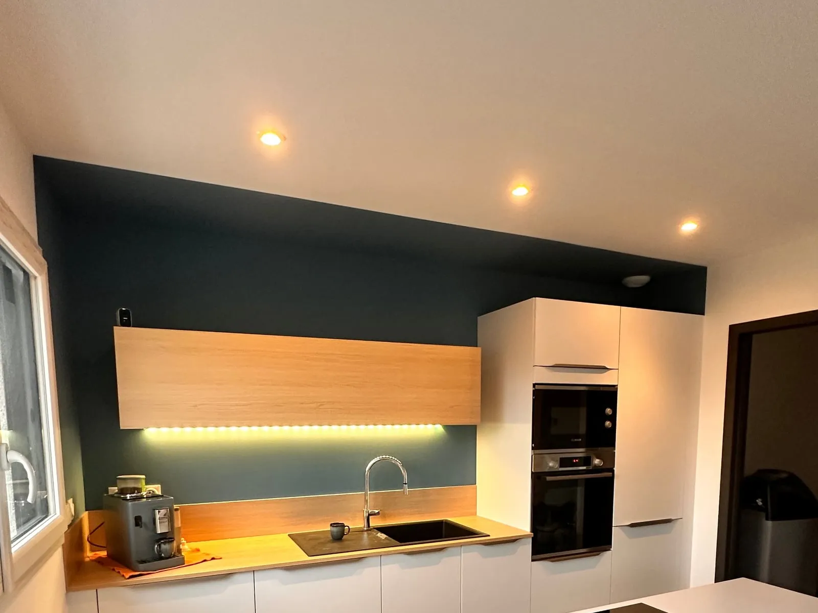 Cuisine avec un mur d’accent peint en bleu pétrole, meubles blancs et façades en bois clair